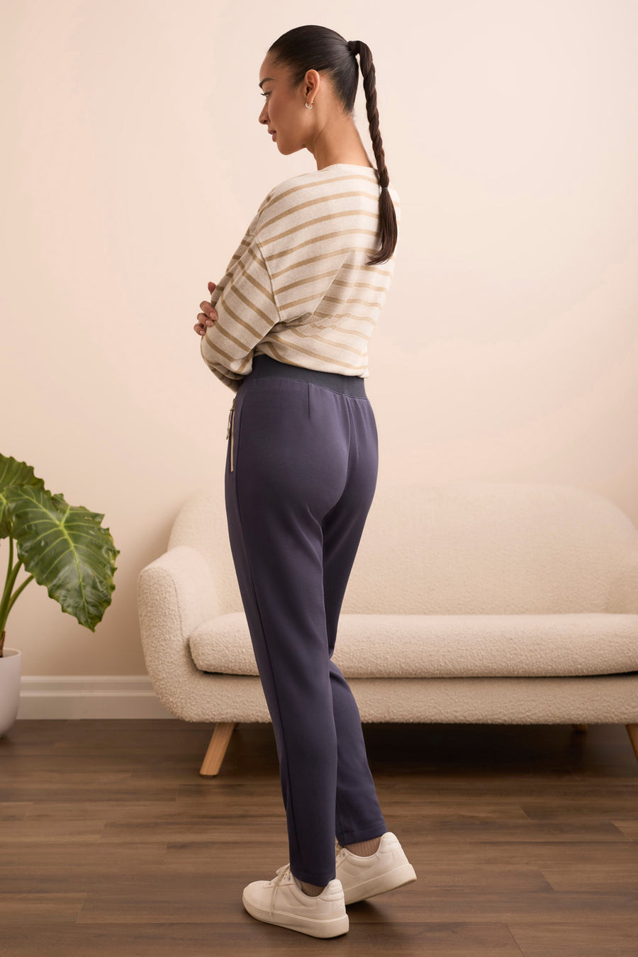 Woman standing with hands on hips wearing a striped sweater and dark pants in a room with a couch and plant.