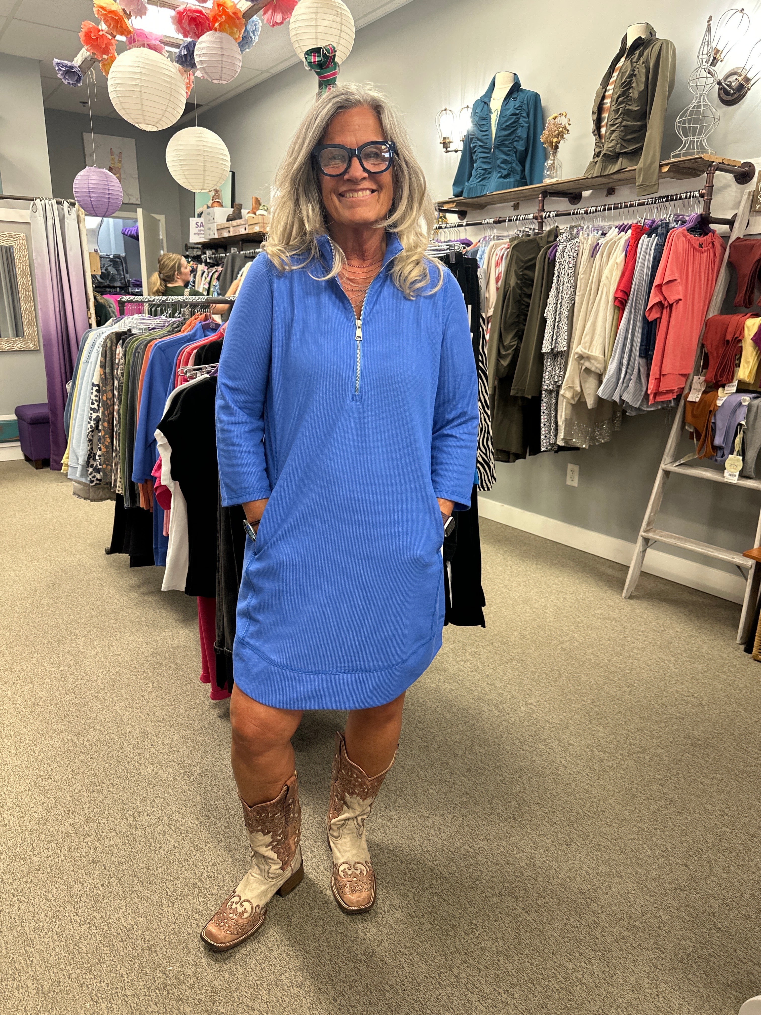 Person wearing a blue sweatshirt and brown boots standing in a store with racks of clothes.