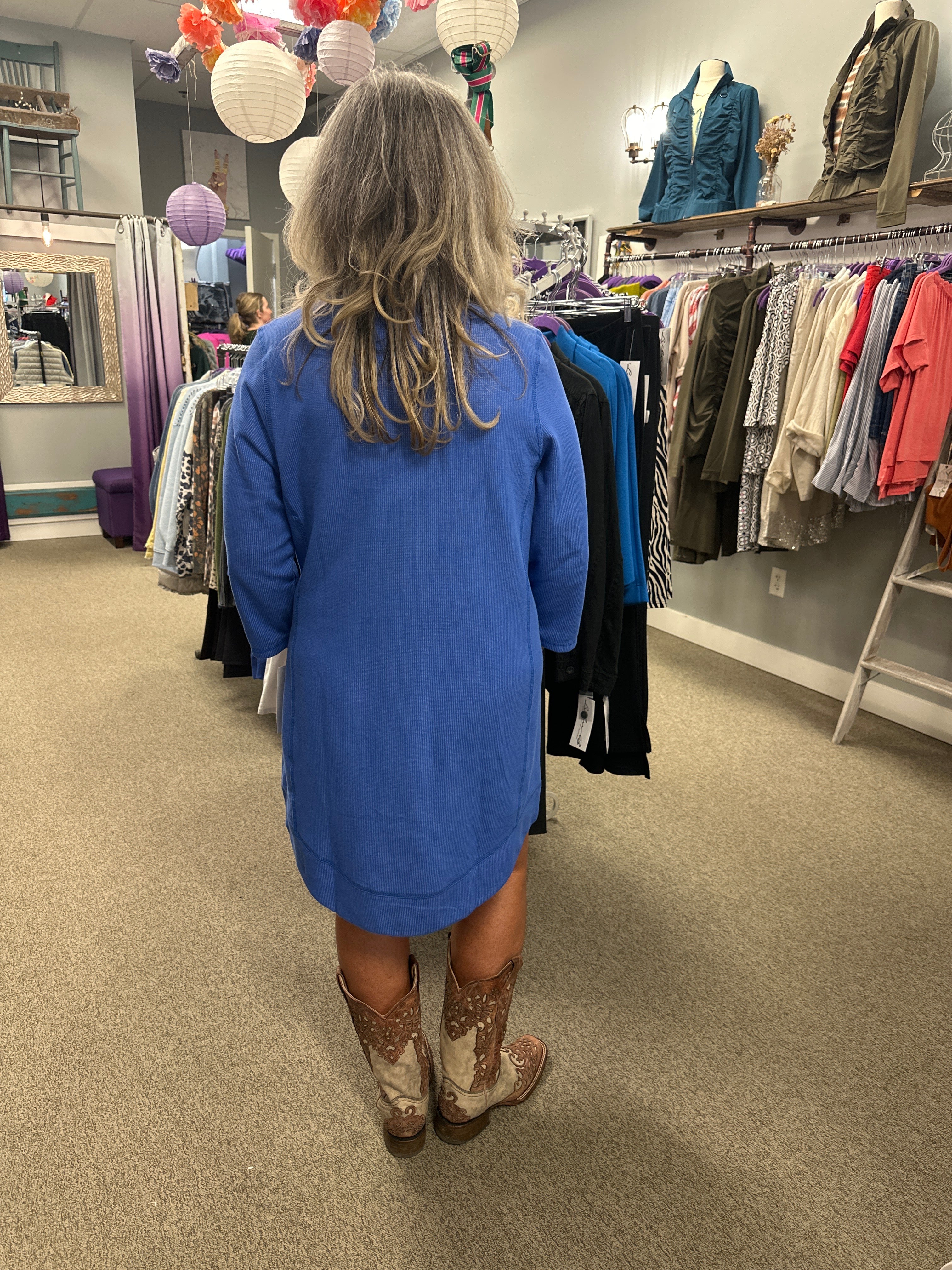 Person wearing a blue dress and cowboy boots standing in a store with racks of clothes.