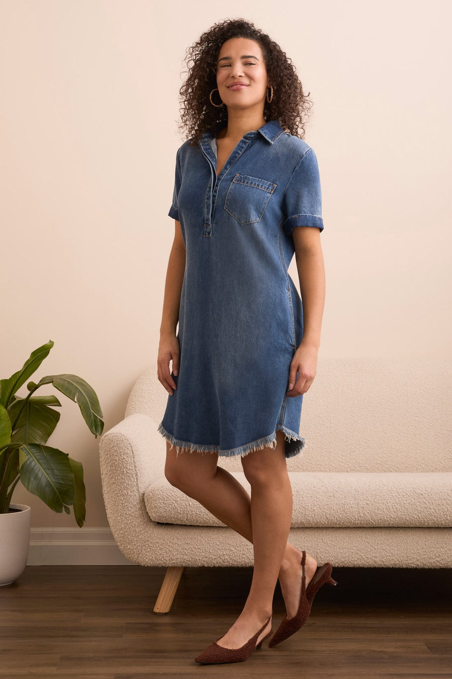 Woman wearing a denim dress standing in a room with a plant and sofa.